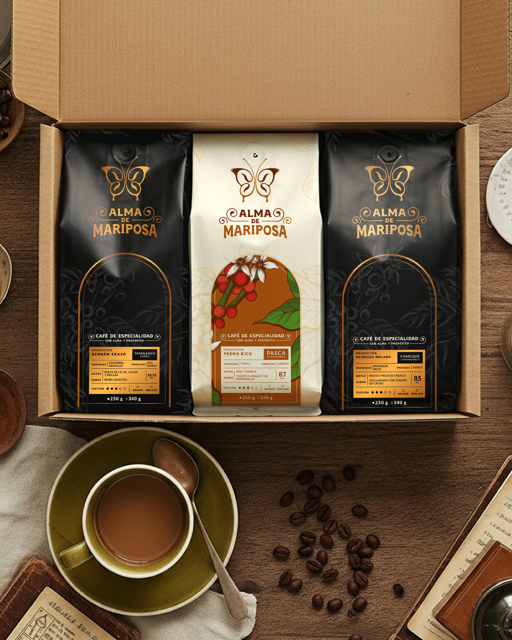 Caja de regalo abierta del Kit Trilogía de Orígenes de Alma de Mariposa. Contiene tres bolsas de café de especialidad de 250g sobre una mesa rústica de madera junto a una taza de café servida.