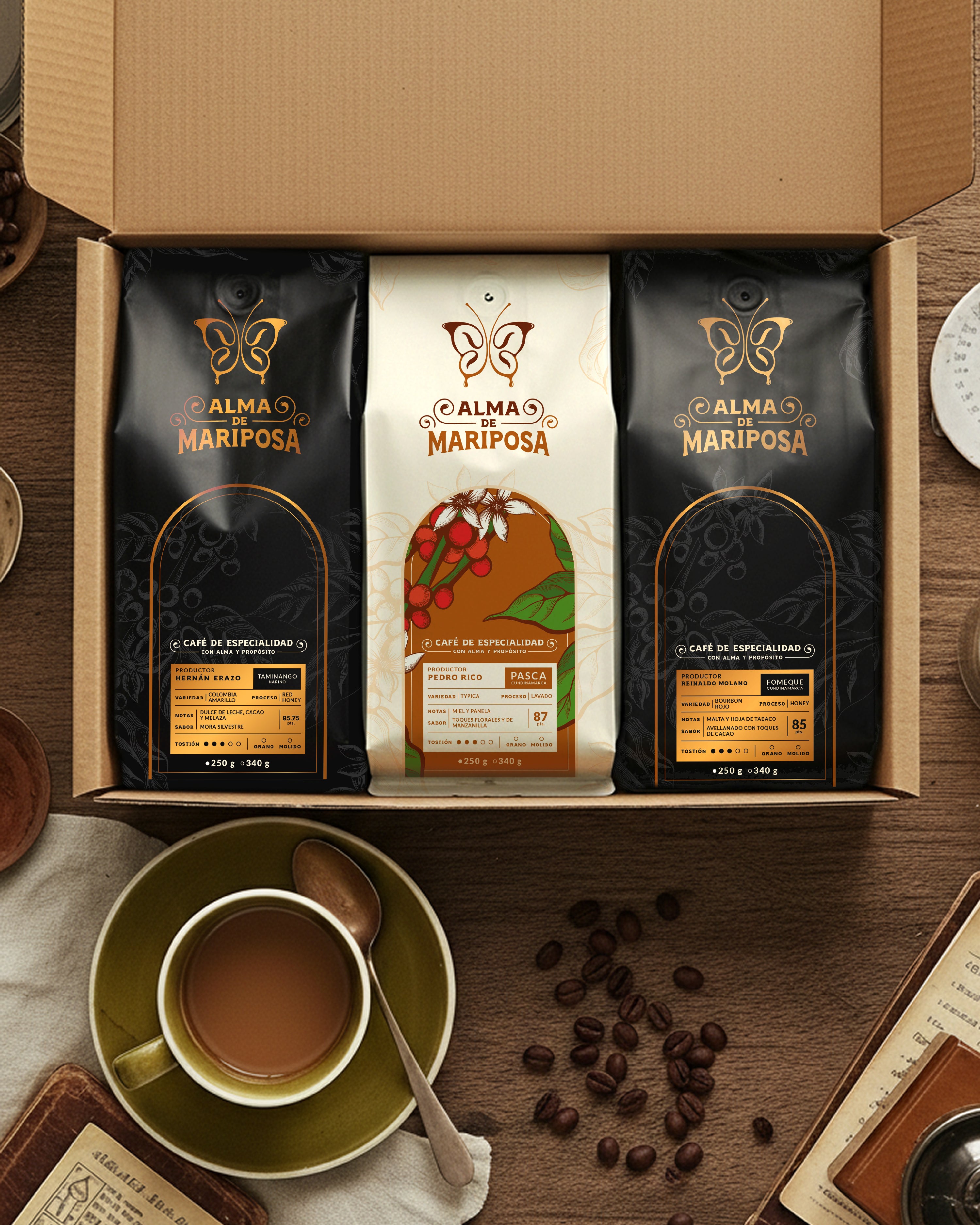 Caja de regalo abierta del Kit Trilogía de Orígenes de Alma de Mariposa. Contiene tres bolsas de café de especialidad de 250g sobre una mesa rústica de madera junto a una taza de café servida.