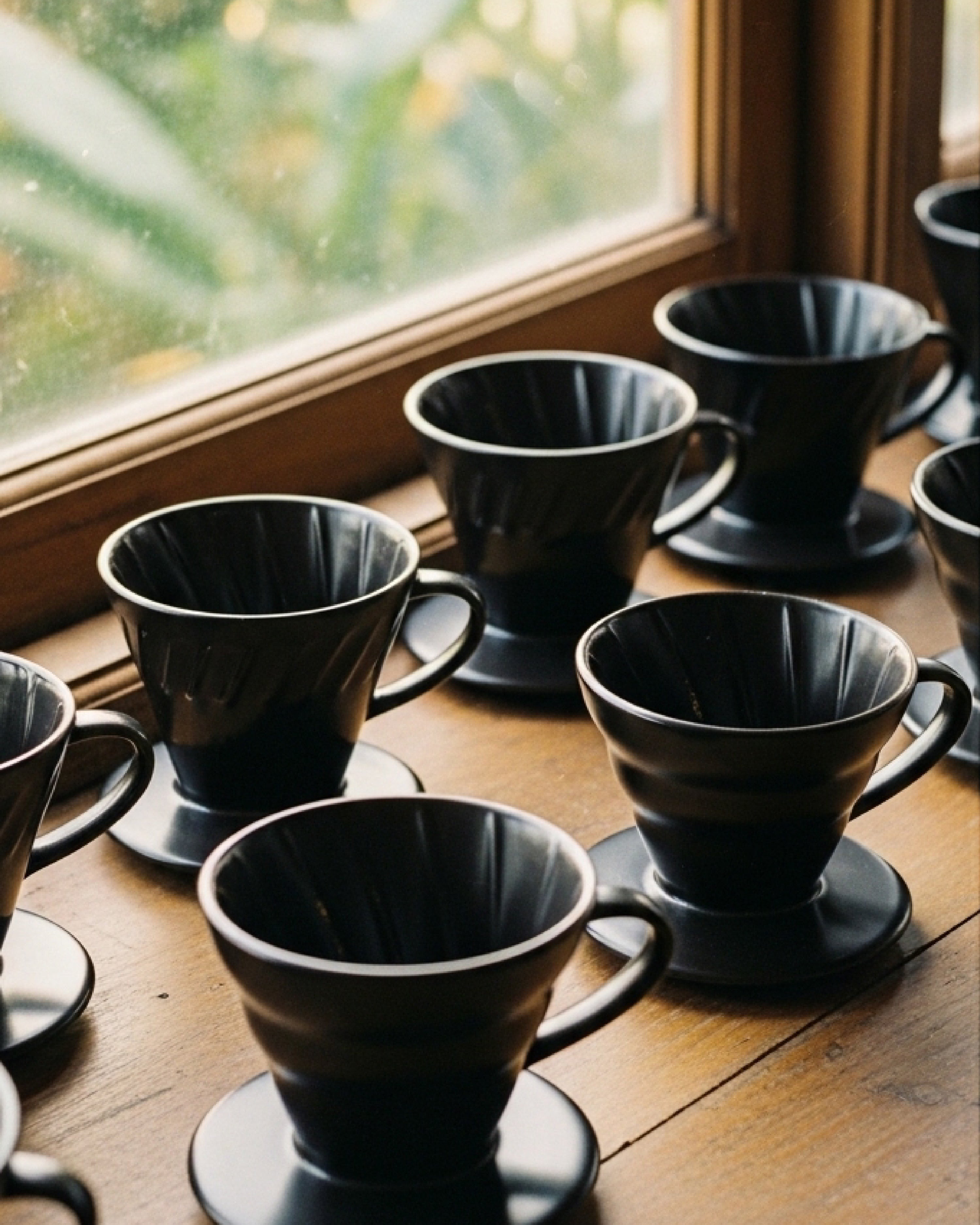 -Colección de Drippers V60 de cerámica negra mate alineados en una repisa de madera junto a una ventana con luz natural.
-Varios métodos de filtrado artesanal para café de especialidad de la marca Alma de Mariposa.