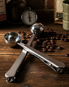 Dos cucharas medidoras de acero inoxidable cruzadas sobre una mesa de madera rústica rodeadas de granos de café tostado. Accesorios para preparar café de especialidad.