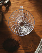 Vista cenital de un dripper V60 de acrílico sobre mesa de madera con café molido. Se aprecian las estrías internas en espiral para una extracción uniforme del café.
