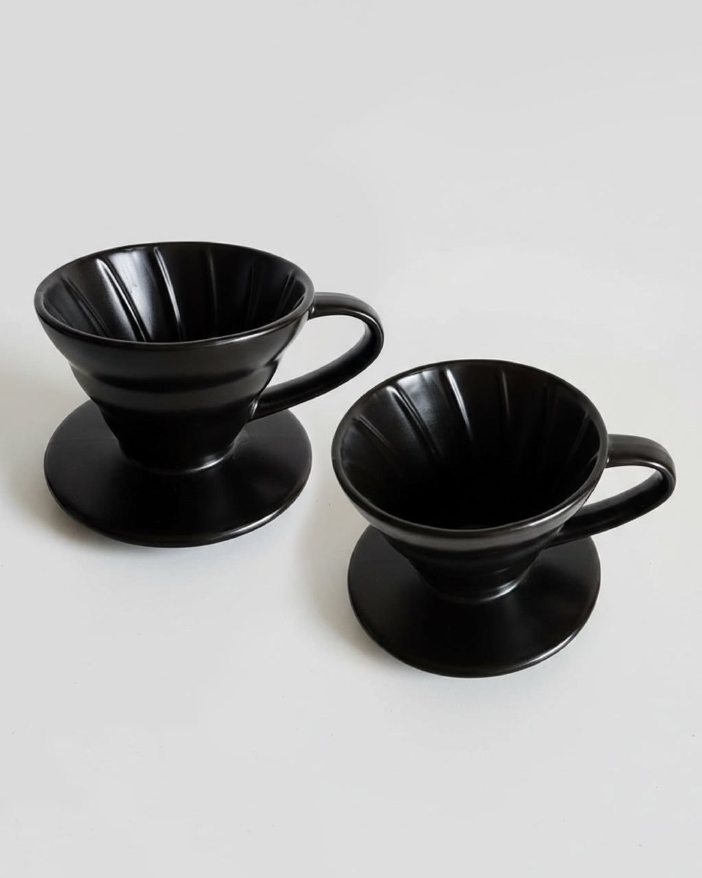 -Vista detallada de dos Drippers V60 de cerámica negra sobre fondo gris claro, mostrando las estrías internas para una extracción uniforme del café.
-Método de goteo profesional en cerámica de alta calidad para Alma de Mariposa.