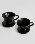 -Vista detallada de dos Drippers V60 de cerámica negra sobre fondo gris claro, mostrando las estrías internas para una extracción uniforme del café.
-Método de goteo profesional en cerámica de alta calidad para Alma de Mariposa.