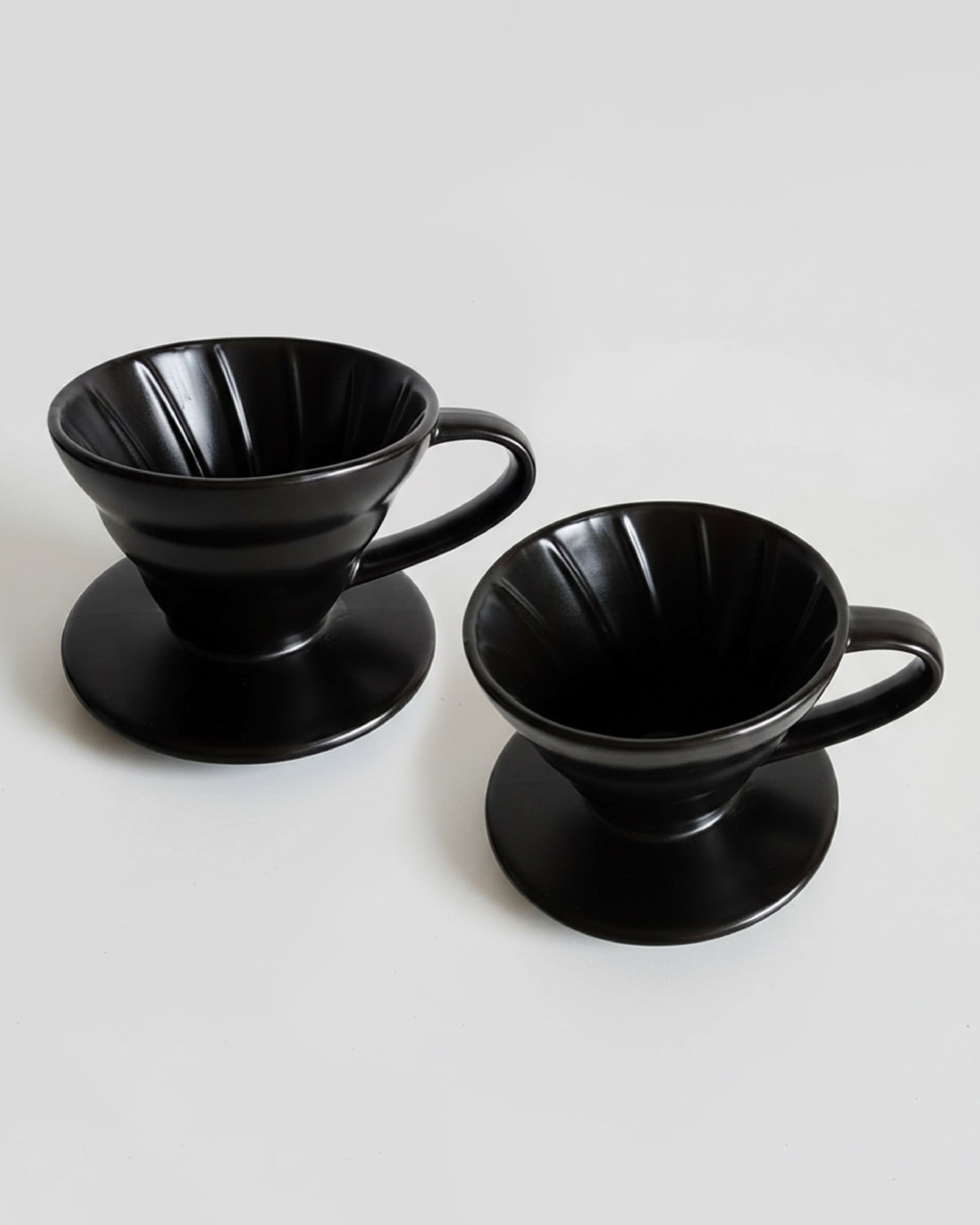 -Vista detallada de dos Drippers V60 de cerámica negra sobre fondo gris claro, mostrando las estrías internas para una extracción uniforme del café.
-Método de goteo profesional en cerámica de alta calidad para Alma de Mariposa.