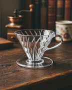 Dripper V60 de acrílico transparente para café filtrado sobre una mesa de madera. Fondo con molino de café manual rústico y libros, resaltando el ritual de Alma de Mariposa.