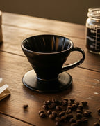 -Dripper V60 de cerámica negra mate sobre una mesa de madera rústica con granos de café esparcidos y luz cálida de atardecer.
-Accesorio profesional para preparar café filtrado en casa con el ritual de Alma de Mariposa.