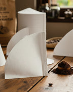Filtro de papel blanco para café V60 en primer plano sobre mesa de madera rústica con una montaña de café recién molido. Accesorio esencial para el ritual de filtrado de Alma de Mariposa.