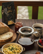 Server de vidrio con café junto a un desayuno de pan artesanal, huevos revueltos y una bolsa de café Alma de Mariposa. Jarra para compartir café de especialidad en casa.