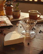 Kit de preparación de café filtrado con Dripper V60 de acrílico y filtros de papel sobre una mesa de madera rústica con granos de café y tazas de la marca Alma de Mariposa.