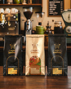 Tres bolsas de café de especialidad Alma de Mariposa de 250g sobre una barra de madera. Kit Trilogía de Orígenes con molino y máquina de espresso de fondo.