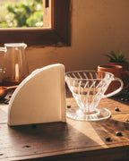Dripper V60 de acrílico transparente junto a un filtro de papel blanco sobre una mesa de madera iluminada por el sol. Al fondo se aprecia un server con café de especialidad.