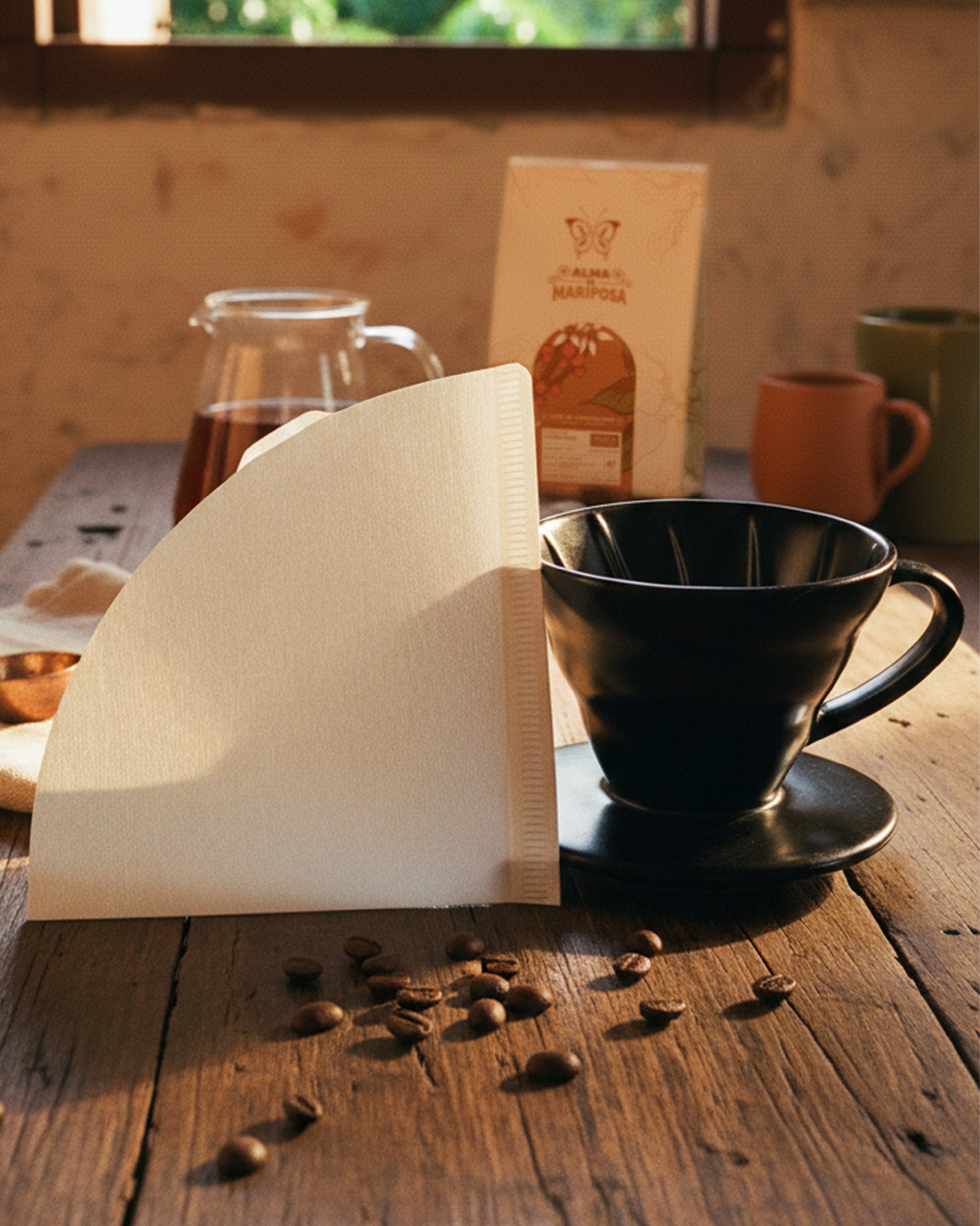 Dripper V60 de cerámica negra junto a un filtro de papel blanco sobre una mesa de madera con luz natural. Al fondo se ve una bolsa de café de especialidad Alma de Mariposa.