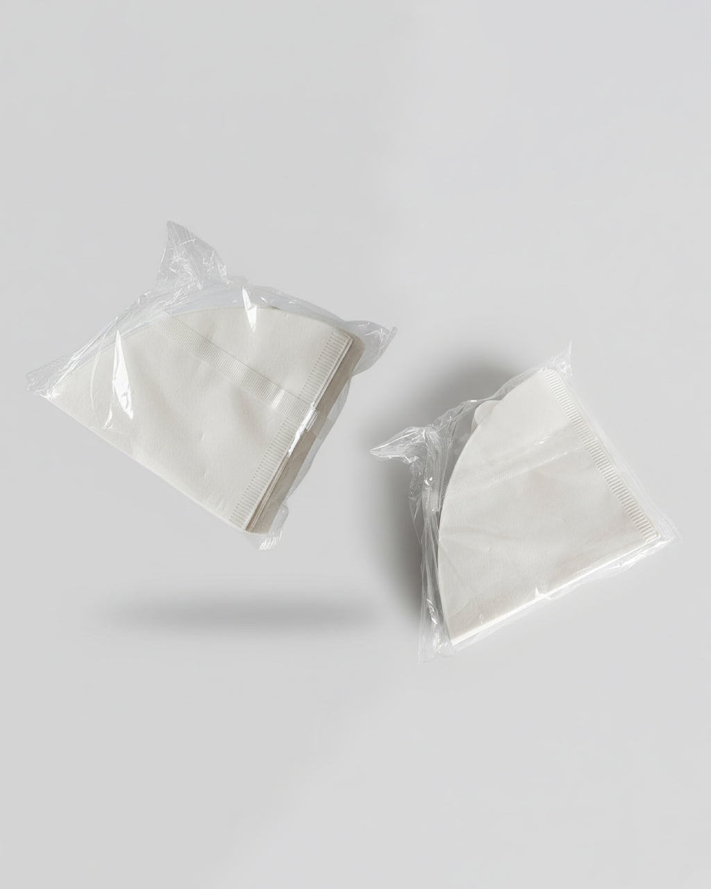 Dos paquetes transparentes de 100 unidades cada uno de filtros de papel blanco para cafetera V60 sobre fondo gris. Repuesto de filtros de alta calidad para café filtrado en Alma de Mariposa.