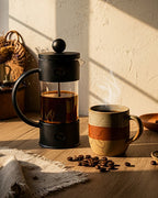 Prensa francesa servida con café caliente y vapor saliendo. Taza de café artesanal sobre mesa de madera con luz de atardecer. Ritual de café en casa.