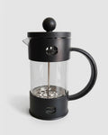 Prensa francesa negra con estructura protectora fija. Cafetera de émbolo con vaso de vidrio y carcasa de plástico resistente. Diseño moderno y seguro.