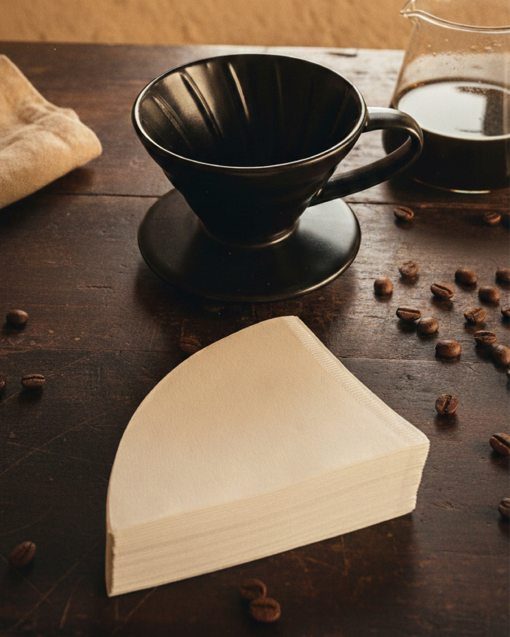 Primer plano de un Dripper V60 de cerámica negra y un paquete de filtros de papel sobre una superficie de madera oscura con granos de café esparcidos.