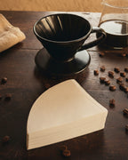 Primer plano de un Dripper V60 de cerámica negra y un paquete de filtros de papel sobre una superficie de madera oscura con granos de café esparcidos.