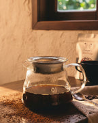 Jarra servidora de vidrio de 360 ml con café de especialidad recién preparado sobre una mesa de madera iluminada por el sol. Accesorio profesional para el ritual de filtrado de Alma de Mariposa.