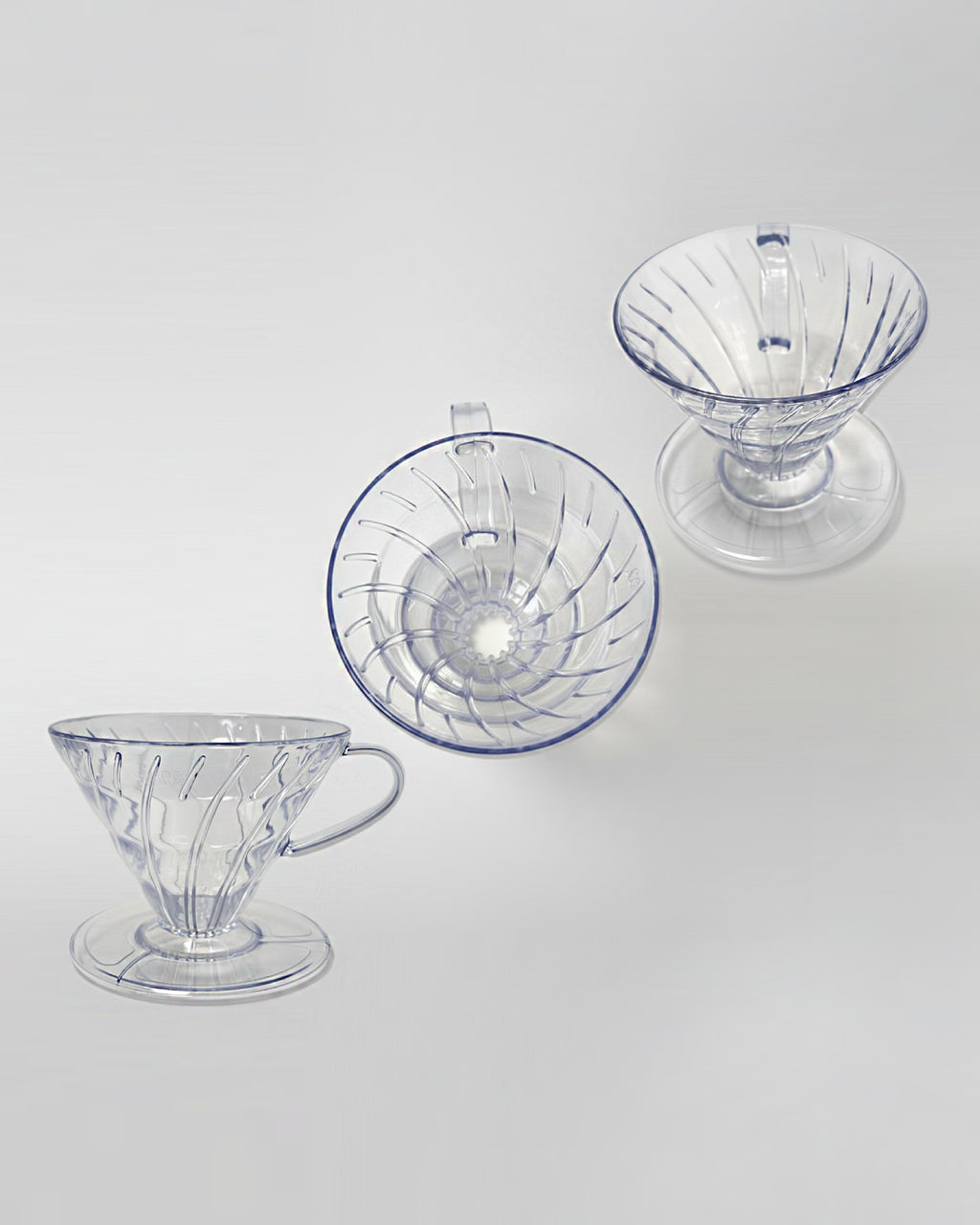 Composición de tres drippers V60 de acrílico transparente vistos desde diferentes ángulos sobre fondo gris claro. Diseño ligero y resistente para café de especialidad.