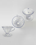 Composición de tres drippers V60 de acrílico transparente vistos desde diferentes ángulos sobre fondo gris claro. Diseño ligero y resistente para café de especialidad.