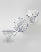 Composición de tres drippers V60 de acrílico transparente vistos desde diferentes ángulos sobre fondo gris claro. Diseño ligero y resistente para café de especialidad.