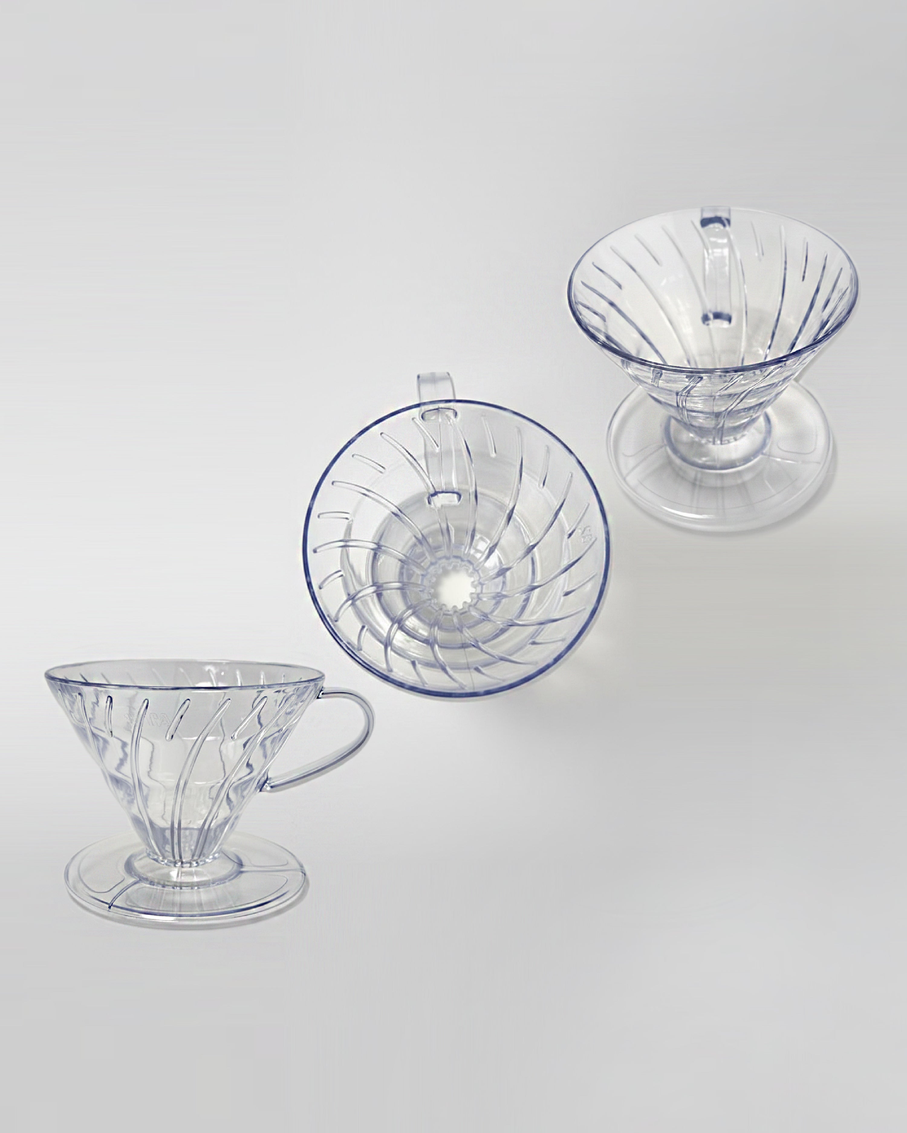 Composición de tres drippers V60 de acrílico transparente vistos desde diferentes ángulos sobre fondo gris claro. Diseño ligero y resistente para café de especialidad.